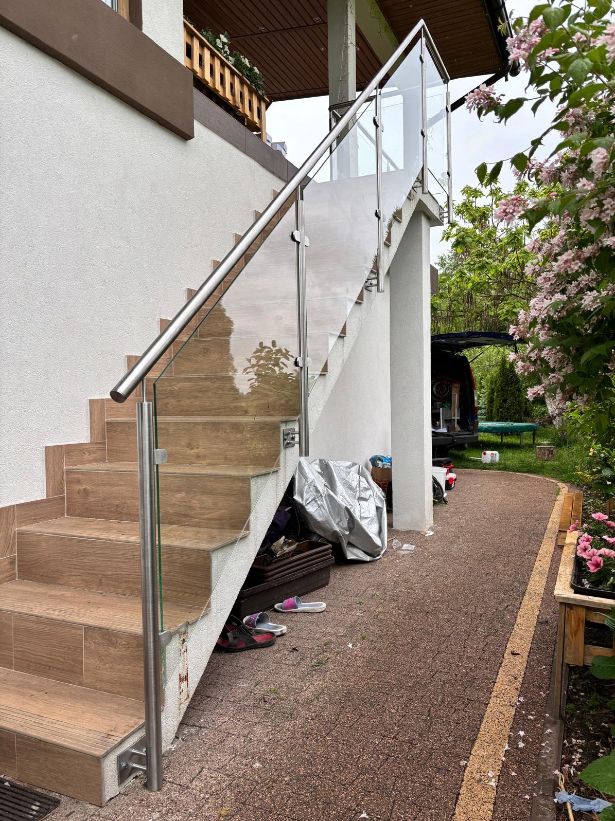 Zewnętrzne schody z drewnopodobnych płytek z balustradą ze szkła i stali nierdzewnej, prowadzące do domu. Widoczne kwiaty i elementy otoczenia.