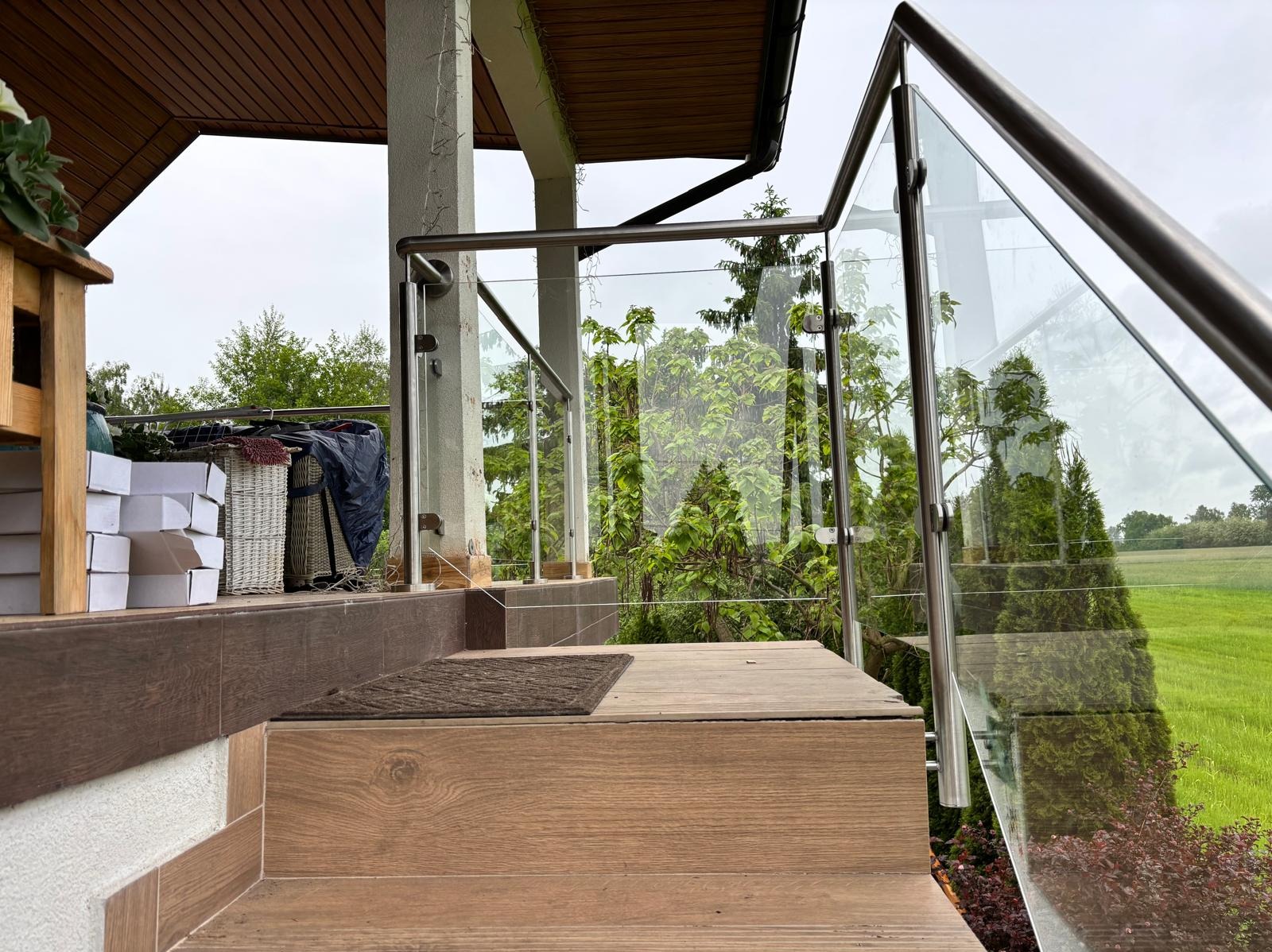 Szklana balustrada z nierdzewnymi elementami mocującymi, zamontowana na schodach prowadzących na taras z drewnianą podłogą. Widok na zieleń ogrodu.