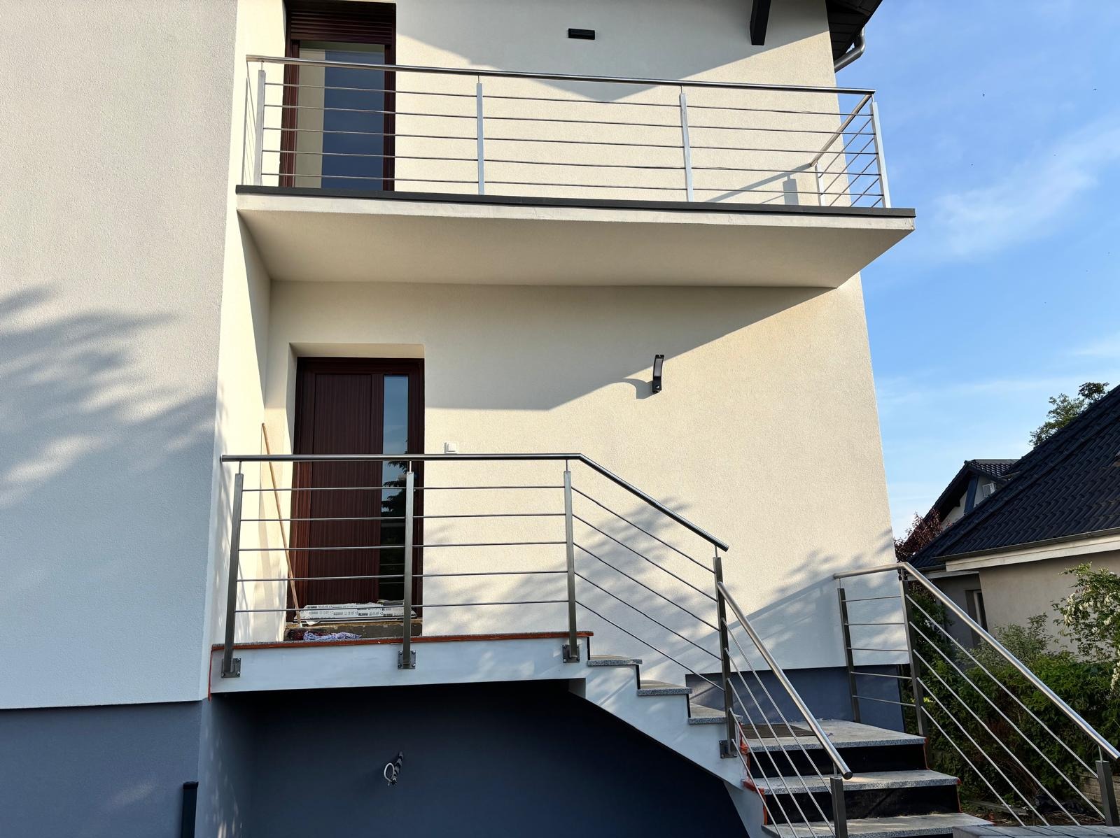 Nowoczesna, wielopoziomowa balustrada ze stali nierdzewnej, z poziomymi prętami, zamontowana przy schodach i balkonie budynku o jasnej elewacji. Widok z dołu.