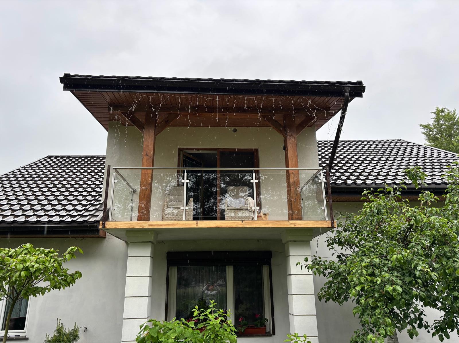 Szklana balustrada na balkonie domu z drewnianą konstrukcją zadaszenia, widok z dołu, deszczowa pogoda.