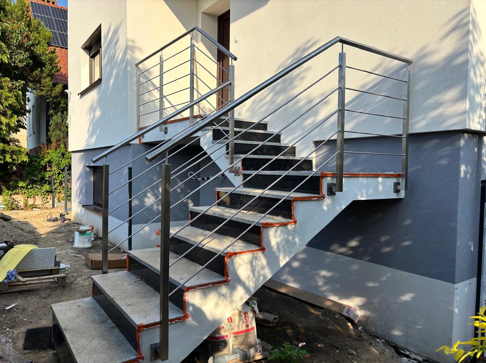 Zewnętrzne schody z czarnymi stopniami i nierdzewną, metalową balustradą, zamontowaną przy jasnym budynku z szarą podmurówką w słoneczny dzień.