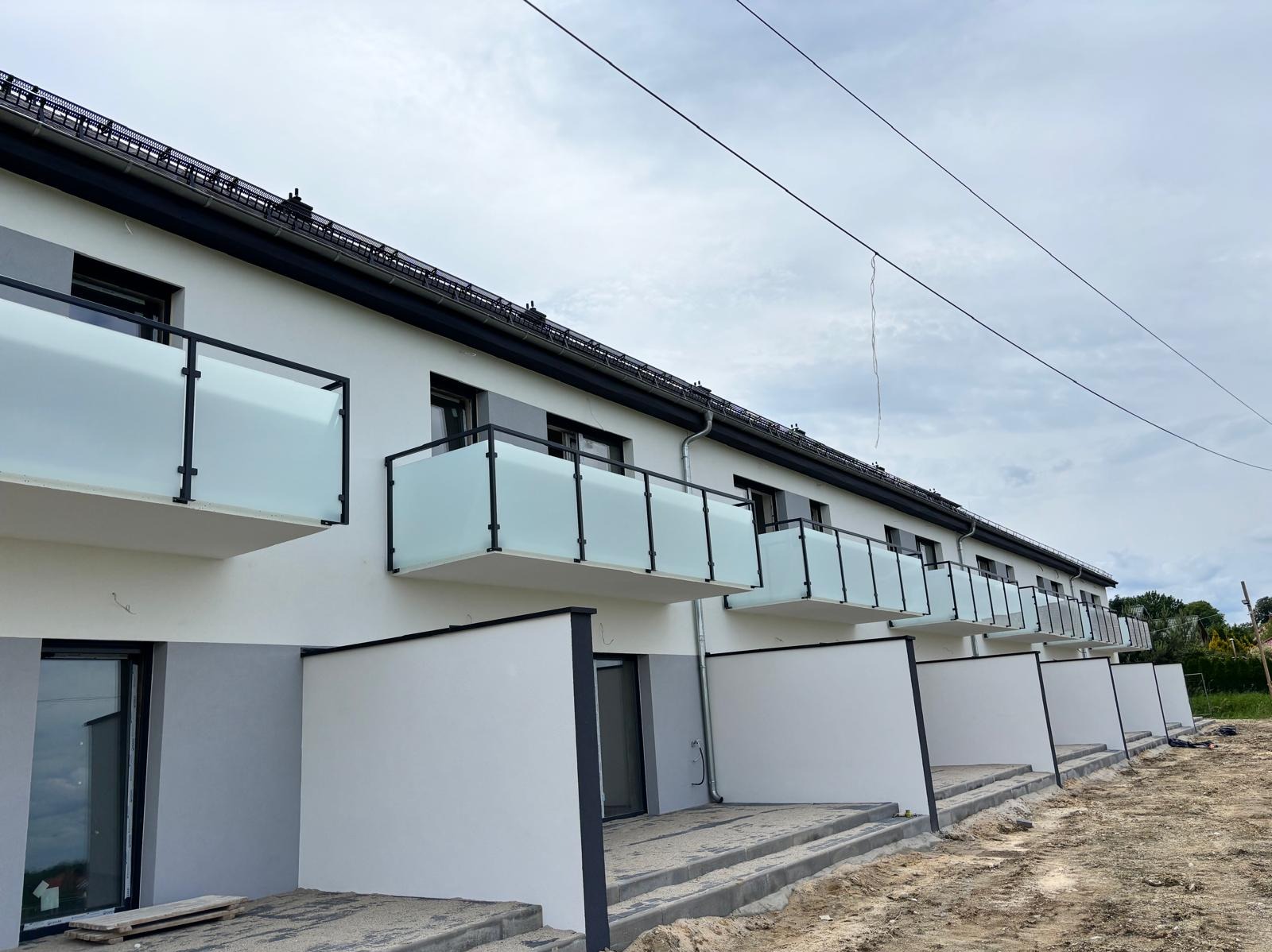 Nowoczesna elewacja budynku z balkonami zabezpieczonymi balustradami ze szkła matowego i czarnymi ramami. Widok z dołu na tle pochmurnego nieba.