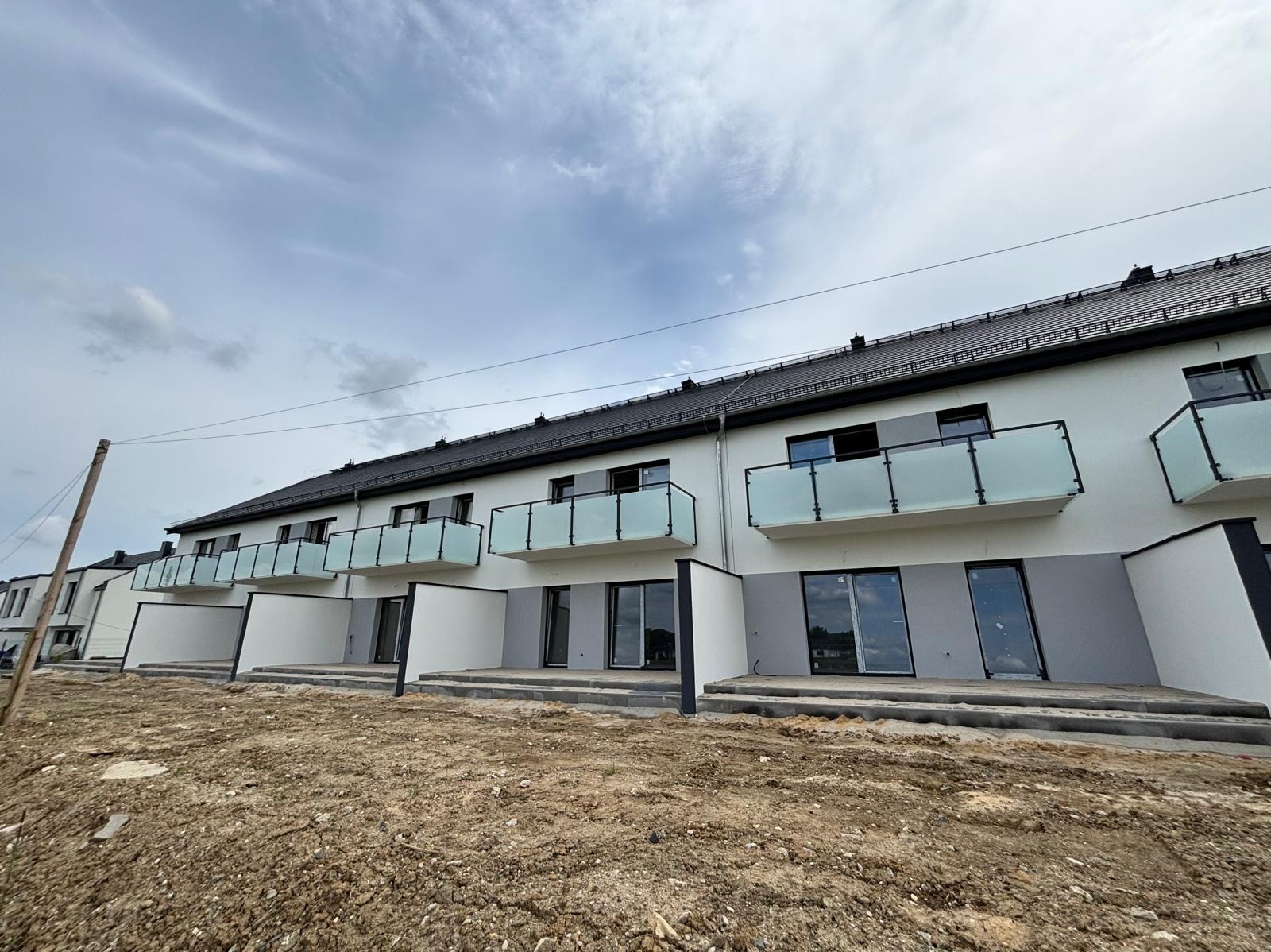 Nowoczesne balkony z mlecznymi szklanymi balustradami w szeregowcu, ziemia przed budynkiem, pochmurne niebo. Minimalistyczny design, świeżo wykończone.