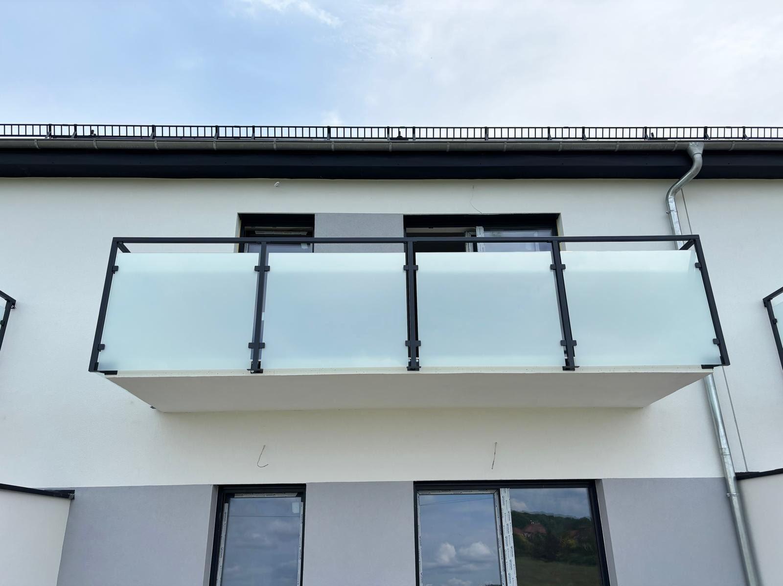 Balkon z matową szybą i czarną ramą, widok z dołu na jasną elewację budynku, proste linie i nowoczesny design.