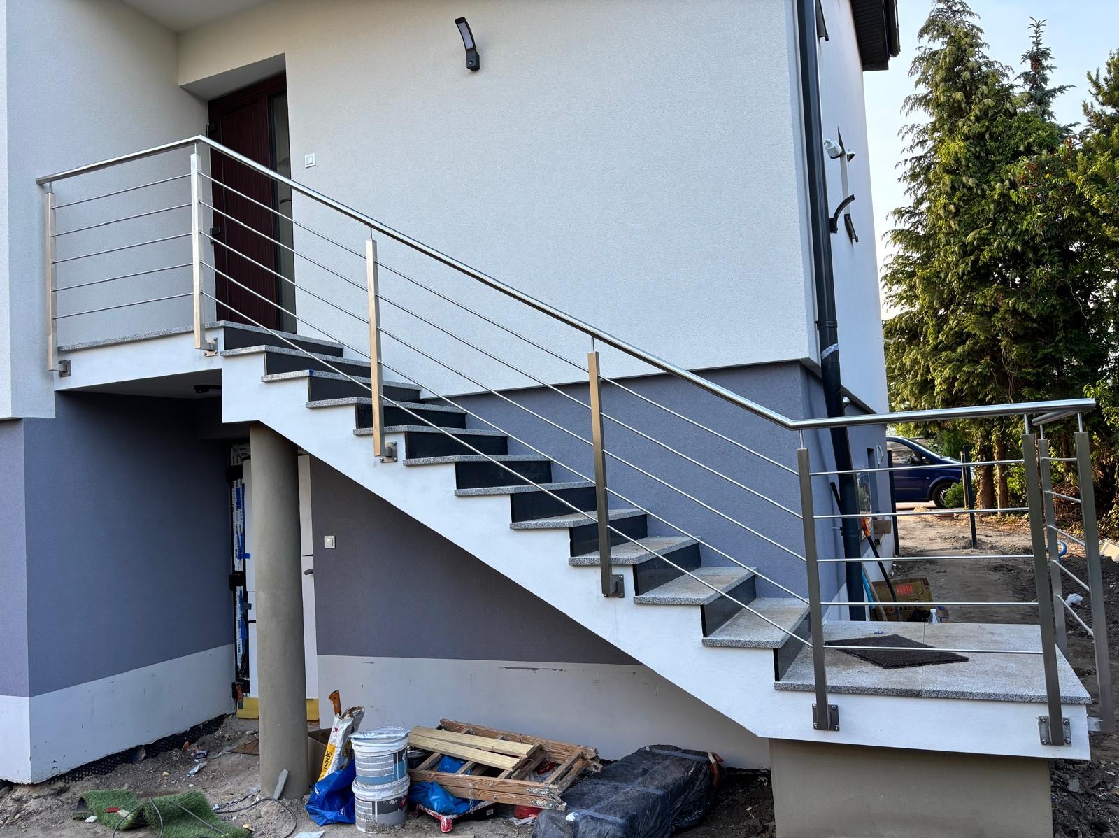 Zewnętrzne schody z poręczą ze stali nierdzewnej przy nowoczesnym budynku. Widoczne detale montażu i wykończenia, otoczenie w trakcie prac budowlanych.