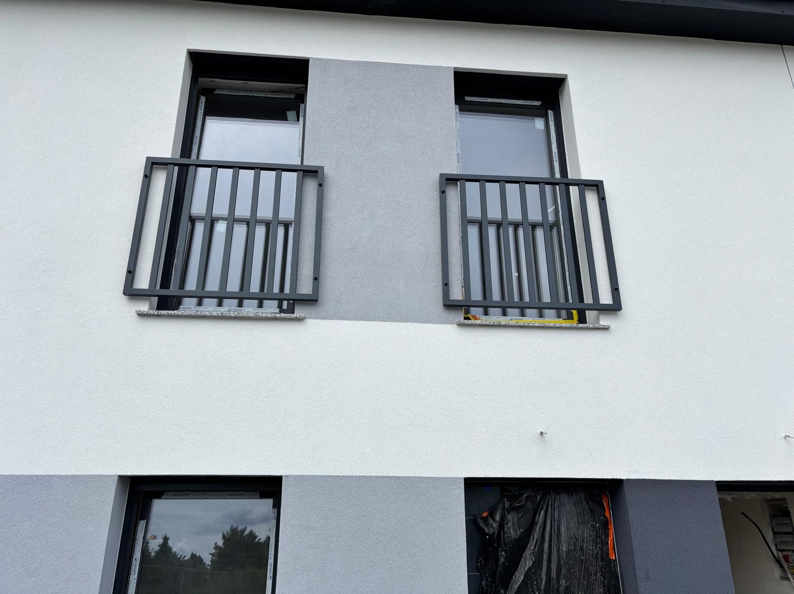 Elewacja budynku z dwoma oknami zabezpieczonymi czarnymi, metalowymi balustradami. Dolne okno zabezpieczone folią. Ściana w odcieniach bieli i szarości.