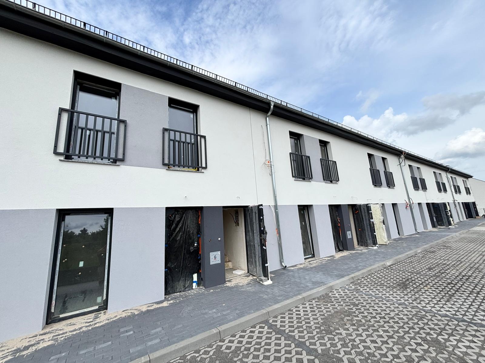 Fasada szeregowca z czarnymi, metalowymi balustradami balkonowymi. Część balkonów zabezpieczona folią. Szara kostka brukowa na chodniku.
