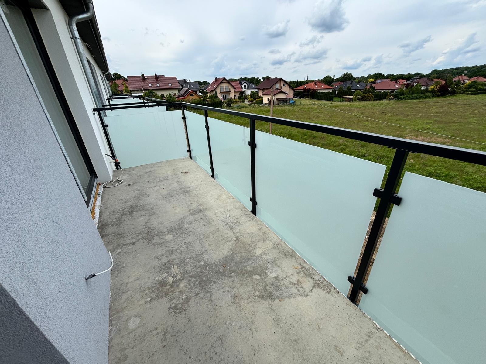 Nowoczesna szklana balustrada balkonowa z czarnymi elementami mocującymi, widok na Żary i okoliczne domy. Minimalistyczny design i funkcjonalność.