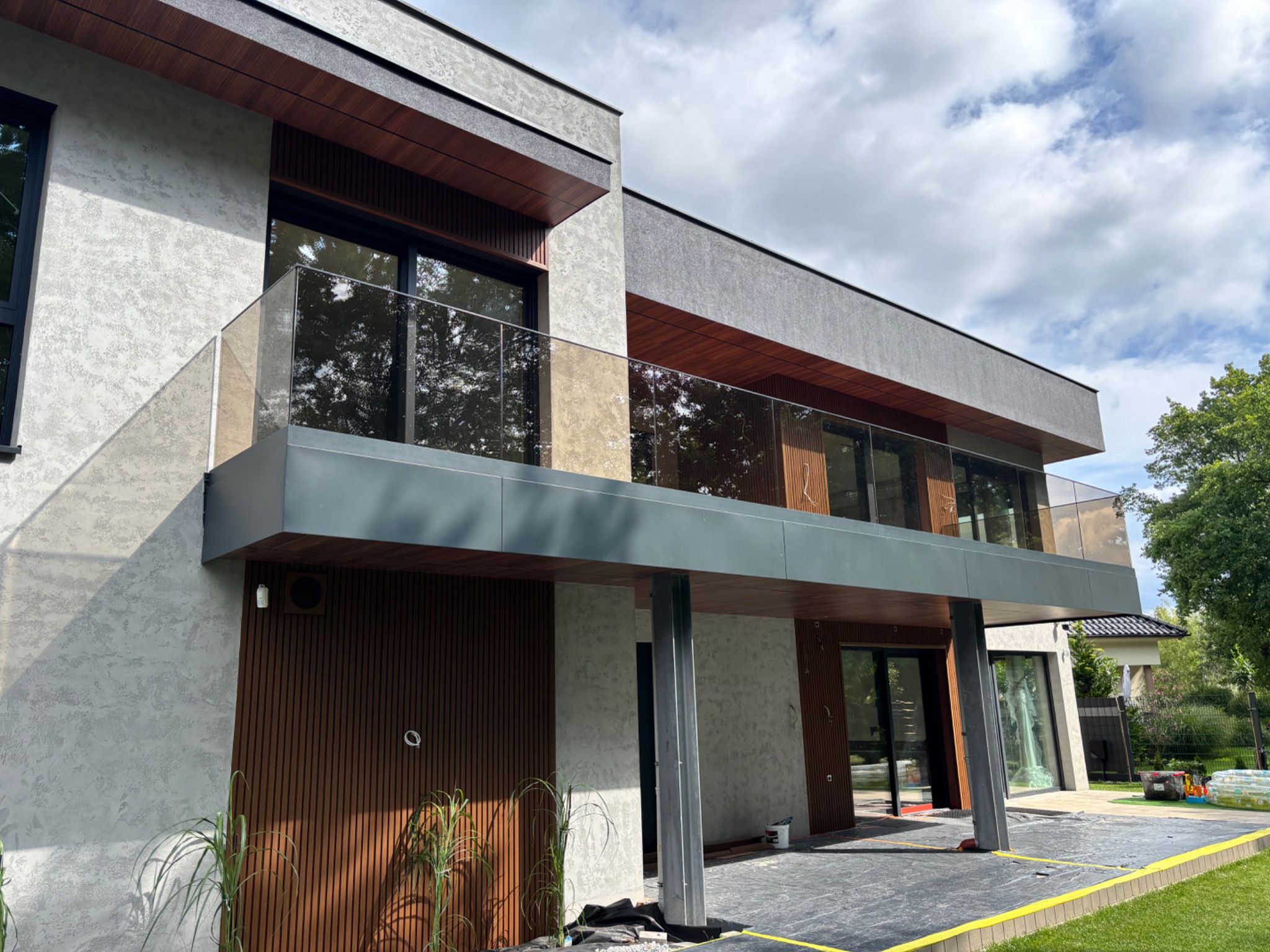 Nowoczesny dom z balkonem i szklaną balustradą. Ujęcie z dołu podkreśla architekturę budynku i detale wykończenia. Widoczne elementy drewniane i szara elewacja.