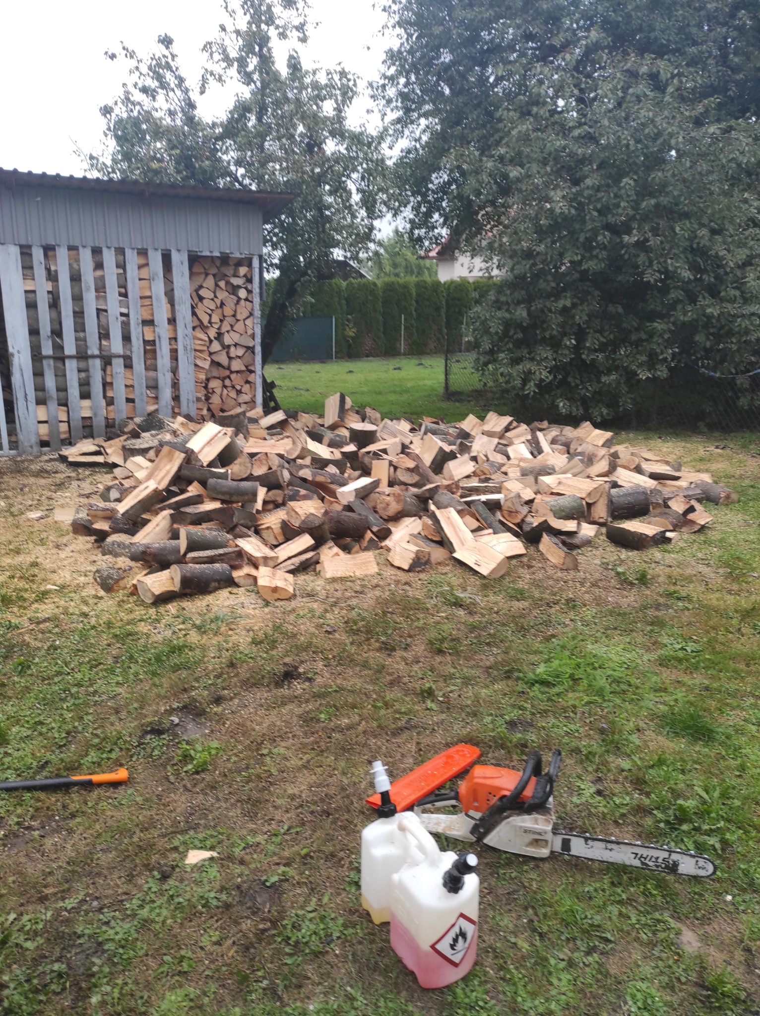 Stos pociętego drewna na trawie obok składu drewna, w tle drzewa i krzewy, w pierwszym planie piła spalinowa i kanister z paliwem oraz siekiera z pomarańczowym trzonkiem.