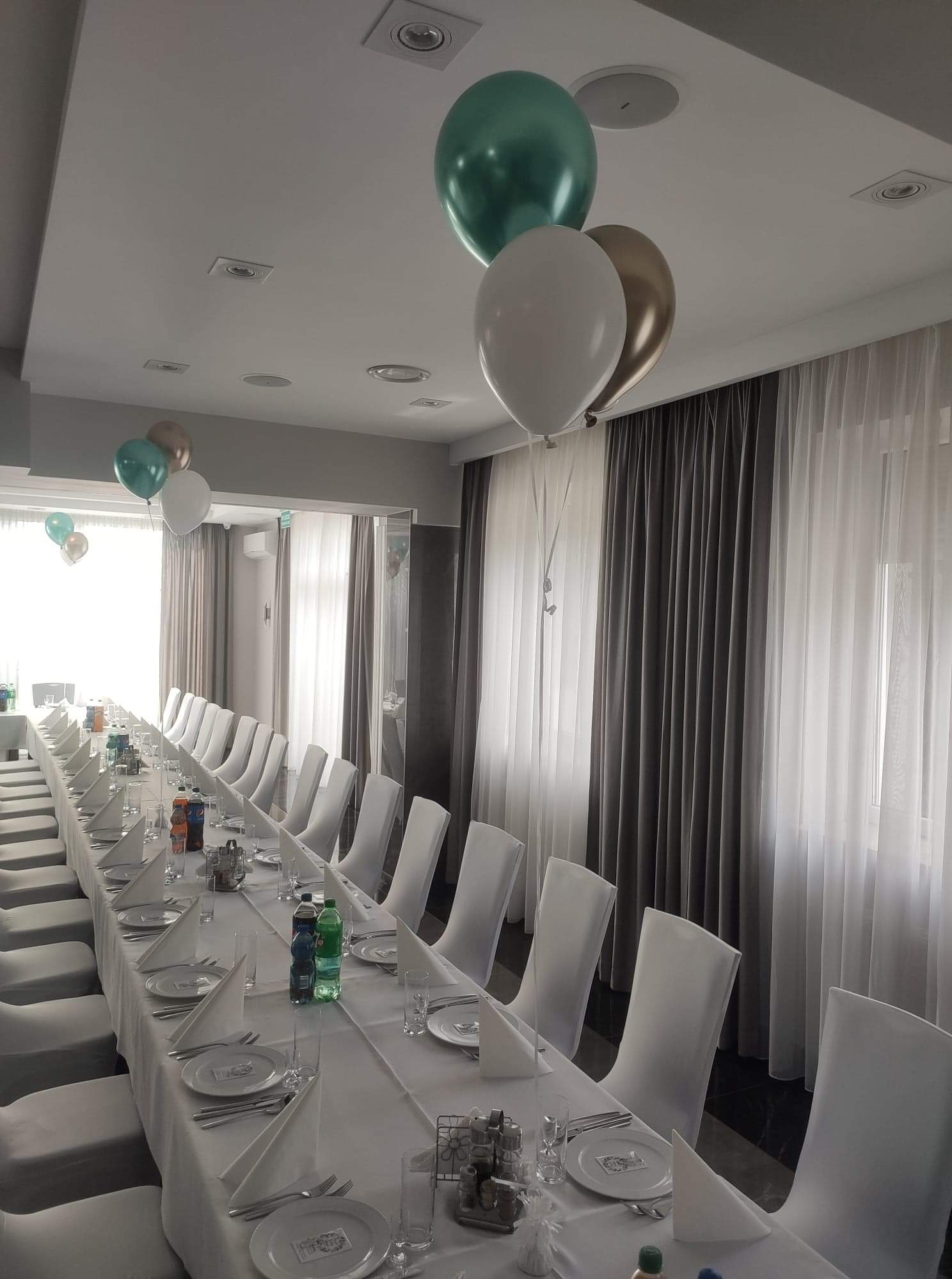 Uroczysta sala z długim stołem nakrytym białym obrusem, eleganckie krzesła w pokrowcach, dekoracyjne balony w odcieniach bieli, złota i zieleni, gotowa na przyjęcie gości.