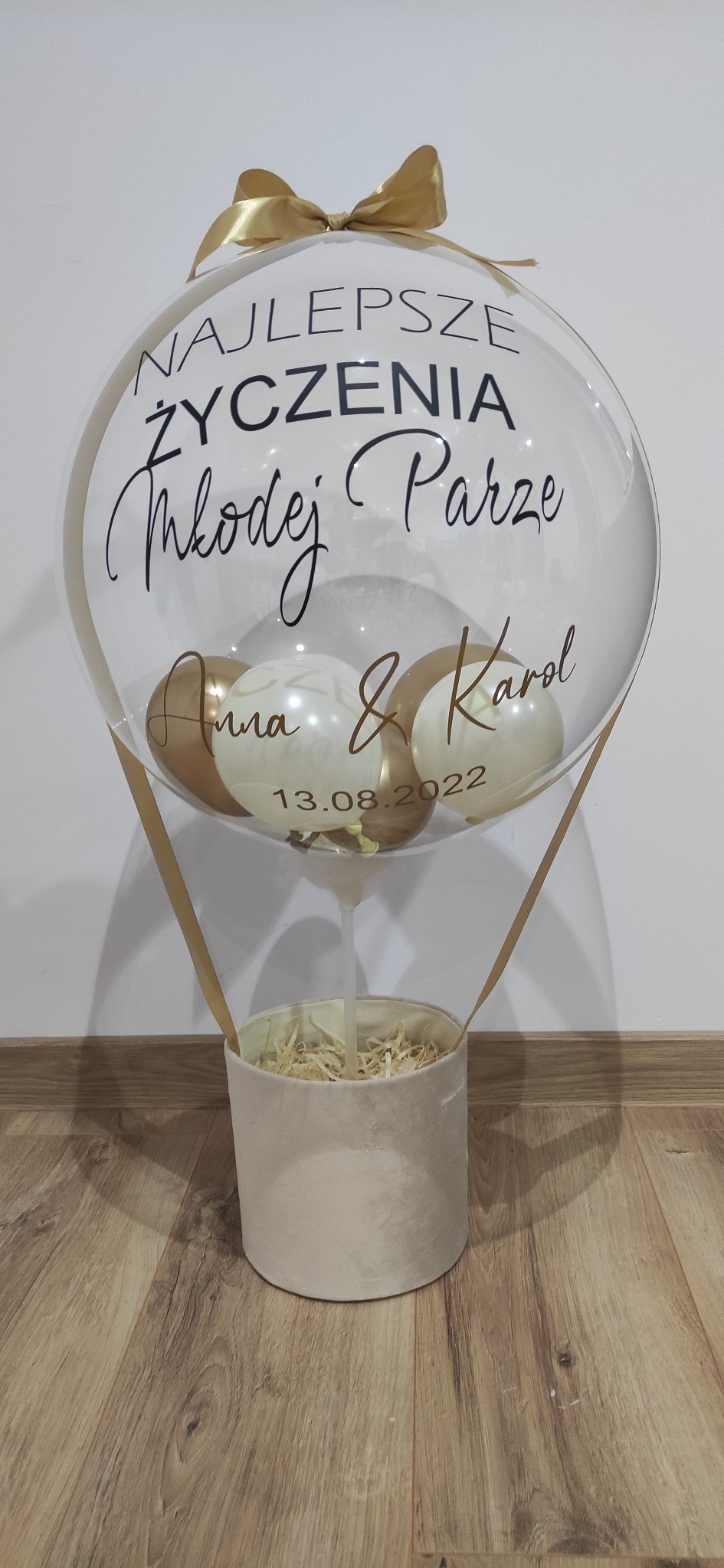Przezroczysty balon z napisem 'Najlepsze Życzenia Młodej Parze', imionami Anna & Karol i datą 13.08.2022, umieszczony w beżowym pudełku z dekoracyjnym wypełnieniem, przewiązany złotą wstążką.
