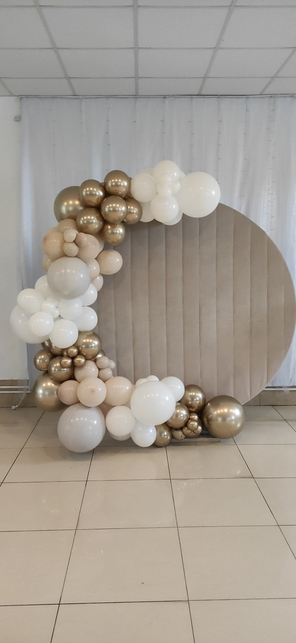 Dekoracja z białych, beżowych i złotych balonów ułożona w łuk przy okrągłej, beżowej ściance na błyszczącej podłodze.