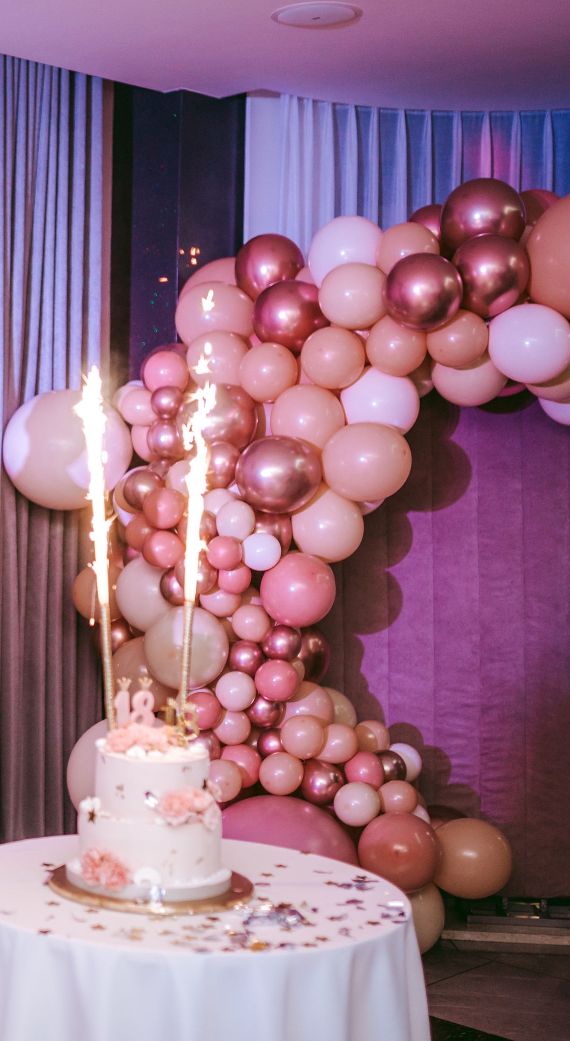 Udekorowany tort urodzinowy z fontannami iskier na stole z białym obrusem, w tle girlanda z różowych, białych i brzoskwiniowych balonów oraz zasłony w odcieniach fioletu.