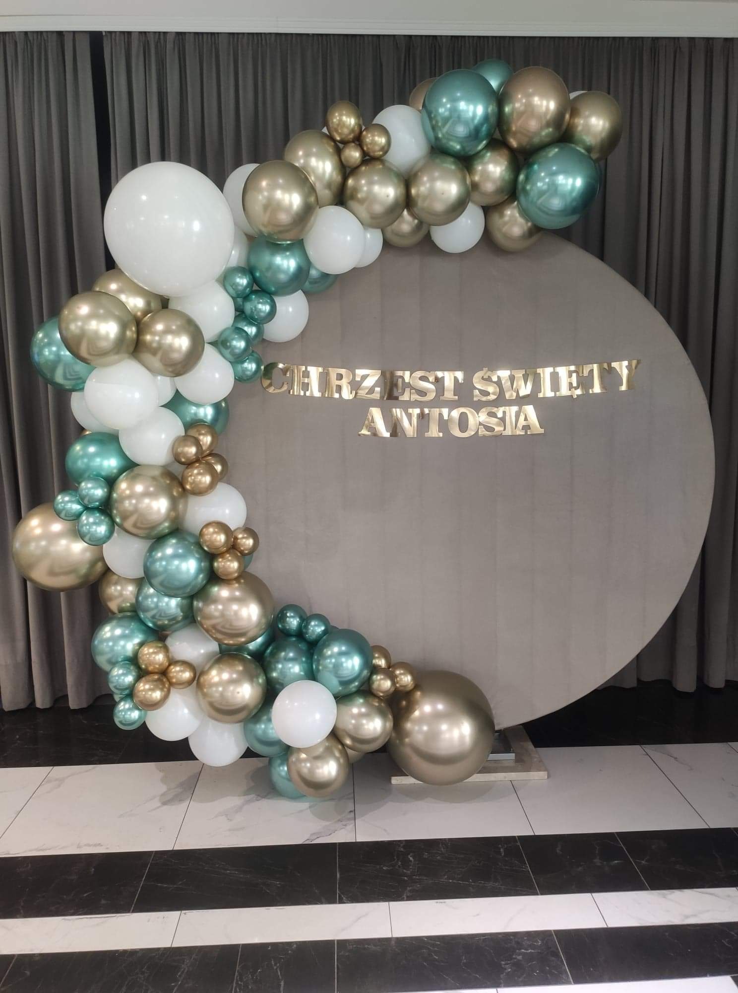 Dekoracja balonowa w kolorach białym, złotym i turkusowym, otaczająca okrągłą ściankę z napisem 'Chrzest Święty Antosia' w złotych literach, na tle szarych zasłon i marmurowej podłogi.