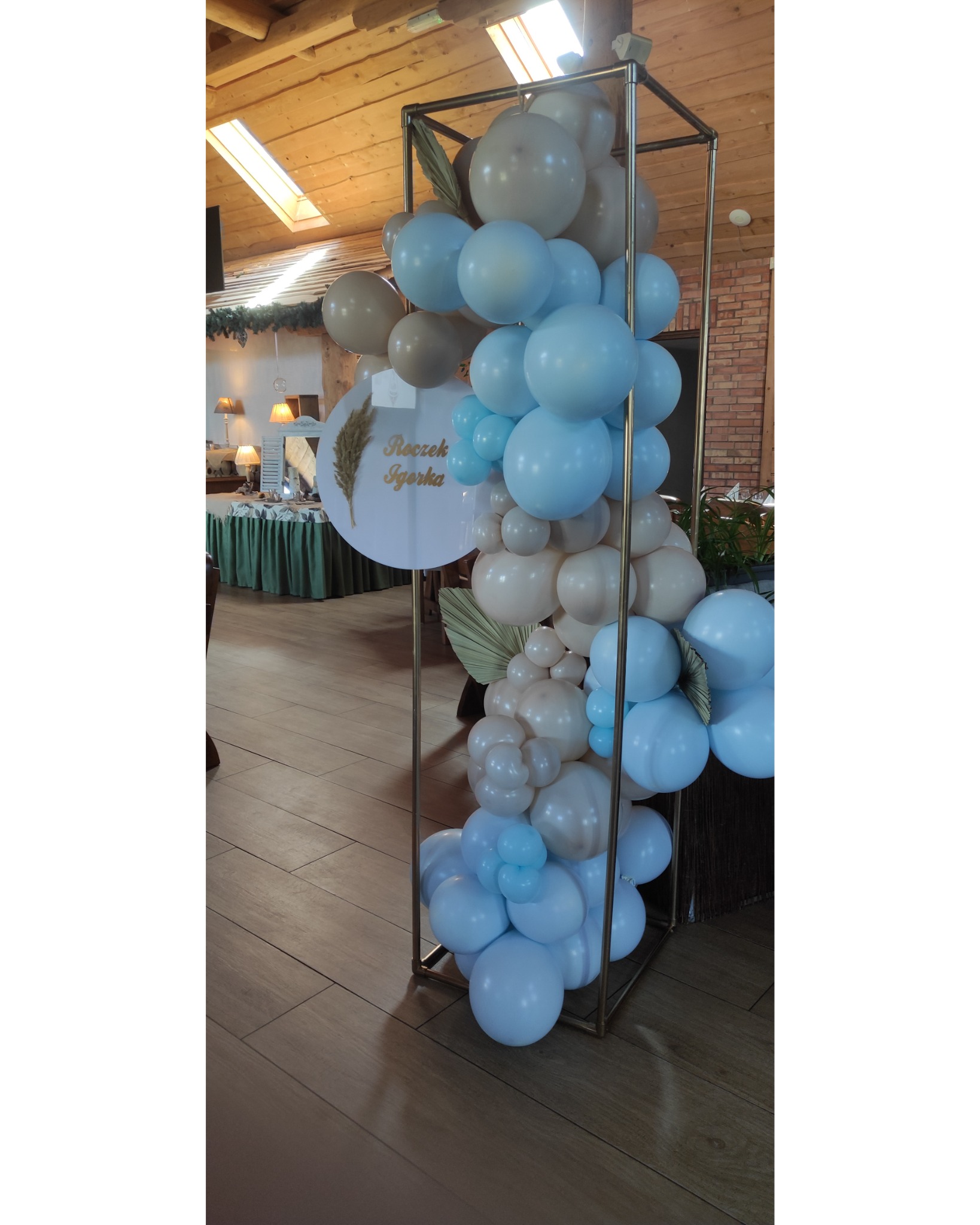 Dekoracja balonowa w kolorach błękitu i beżu, umieszczona w metalowej ramie, z okrągłą tabliczką z napisem 'Roczek Igorka', ustawiona w sali z drewnianym sufitem i oknami dachowymi.