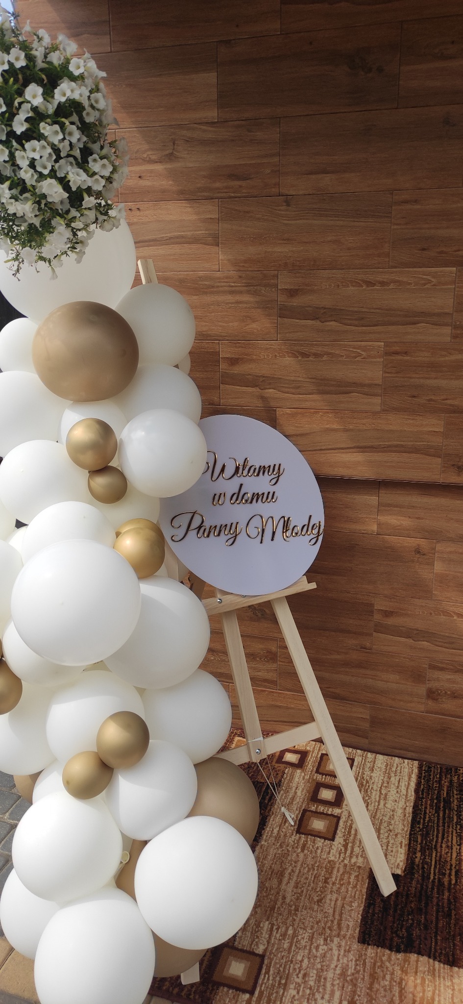Dekoracja wejścia na uroczystość ślubną: białe i złote balony, kwiaty w donicy oraz tablica powitalna na sztaludze z napisem 'Witamy w domu Panny Młodej'.