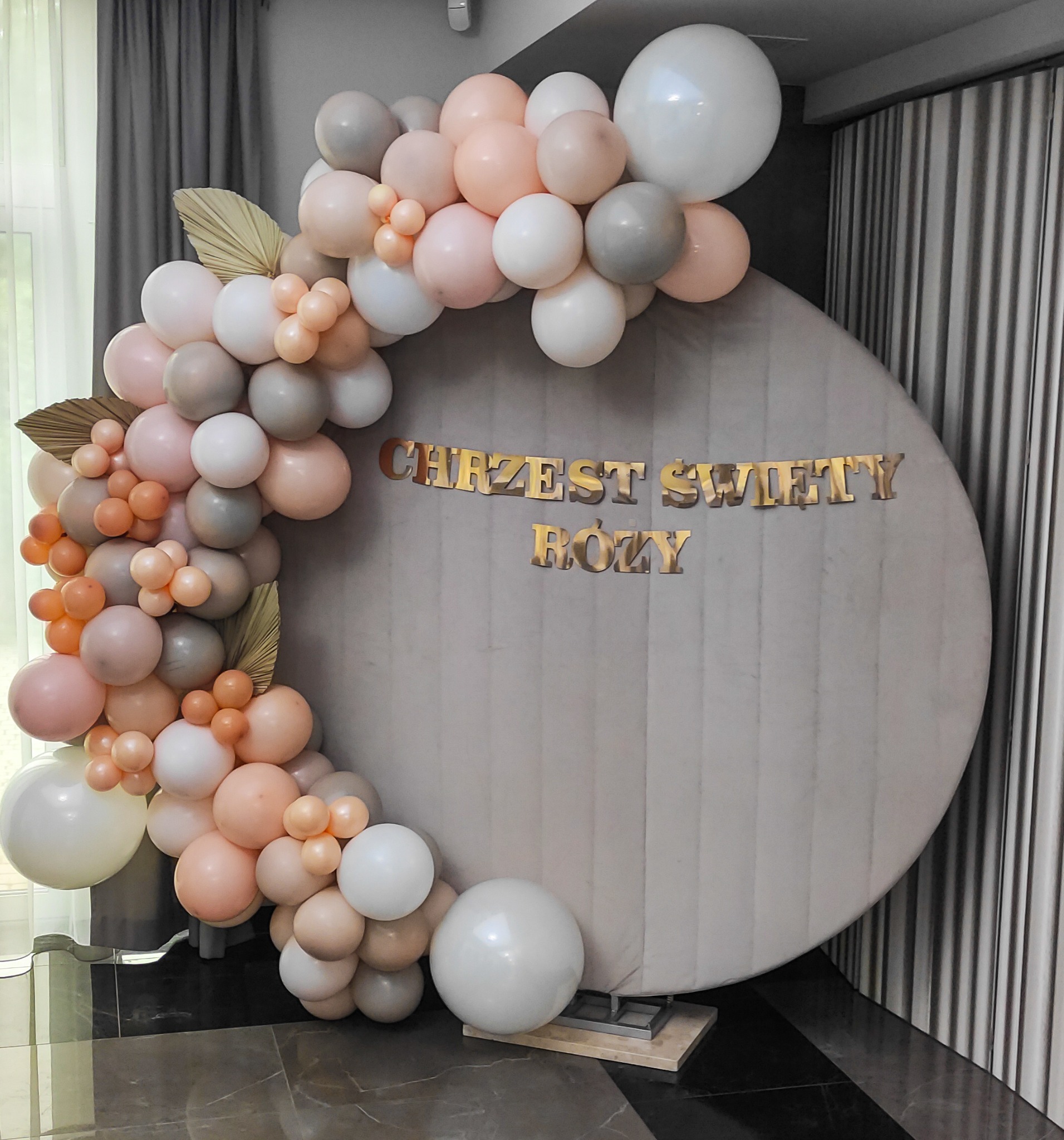 Dekoracja balonowa w odcieniach bieli, szarości i brzoskwini na Chrzest Święty Róży, z suszonymi liśćmi palmowymi, na tle okrągłej, szarej ścianki i ciemnej podłogi.