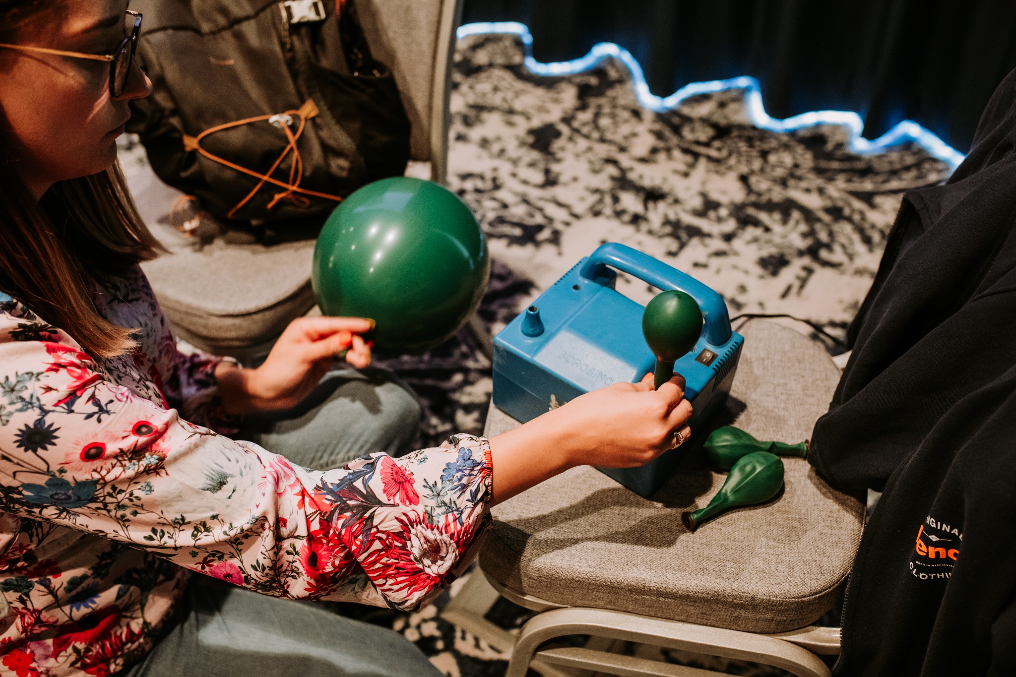 Osoba w kwiecistej bluzce pompuje zielone balony za pomocą niebieskiej pompki elektrycznej w Troszynie, przygotowując się do dekoracji.