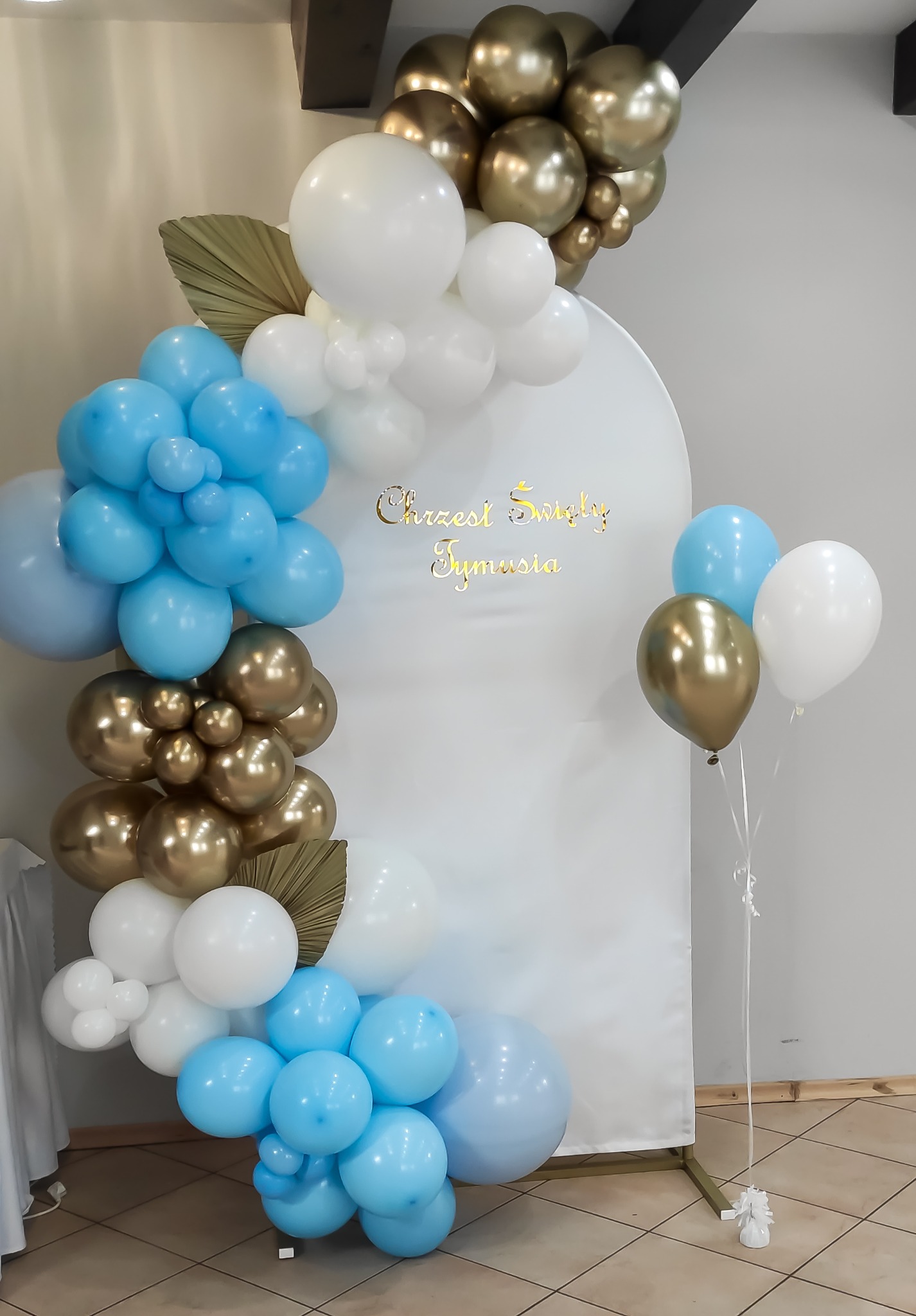 Dekoracja balonowa w odcieniach błękitu, złota i bieli z napisem 'Chrzest Święty Tymusia' na białym tle, uzupełniona o suszone liście palmowe.