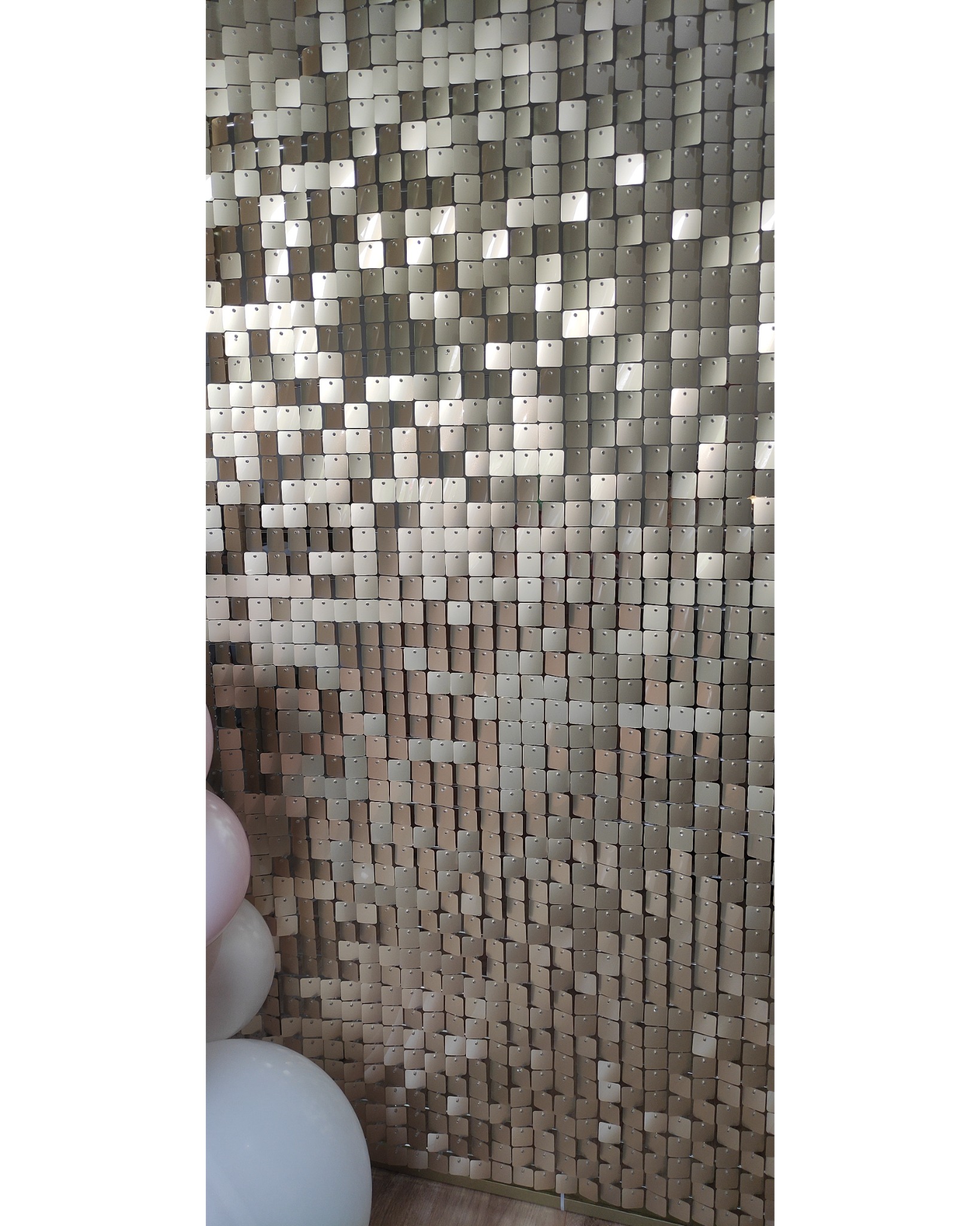 Błyszcząca ścianka dekoracyjna z kwadratowych, ruchomych płytek w odcieniach złota i srebra, z fragmentem białego i różowego balonu po lewej stronie.