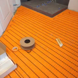 Pomarańczowa mata grzewcza podłogowa ułożona w łazience, widoczny fragment brodzika z szarą mozaiką na ścianie, rolka z matą, pudełko z akcesoriami i taśma klejąca.