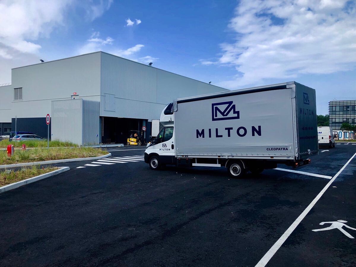 Ciężarówka firmy Milton z naczepą w kolorze szarym, zaparkowana przed magazynem z otwartą bramą, w której widoczny jest wózek widłowy.