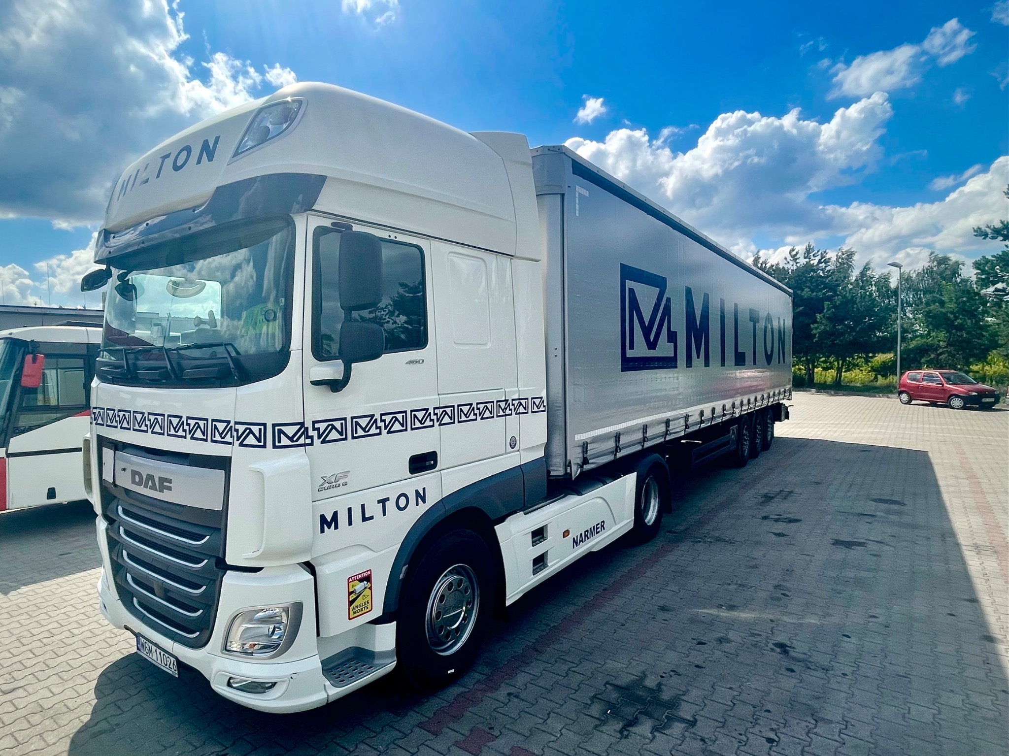 Biała ciężarówka DAF z naczepą z logo firmy Milton na parkingu w słoneczny dzień. Na naczepie widoczny napis Milton. Na pierwszym planie kostka brukowa.