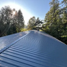 Błękitny dach z blachy trapezowej, widok z góry na tle zieleni drzew i błękitnego nieba z promieniami słońca. Widoczne łączenia blach.