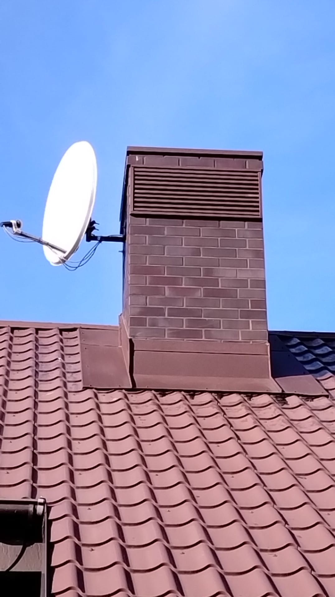Ceglany komin z anteną satelitarną na brązowym dachu z dachówki ceramicznej, na tle błękitnego nieba. Komin posiada kratkę wentylacyjną w górnej części.