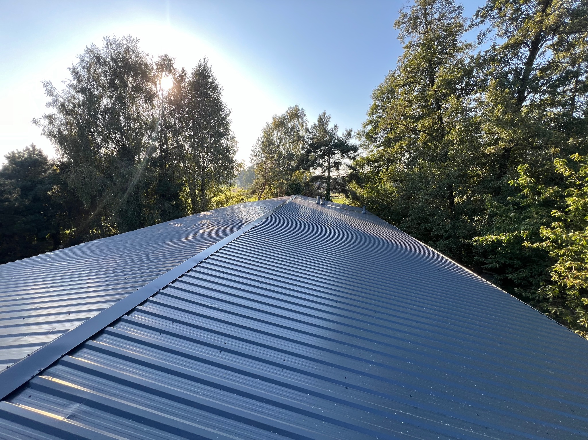 Błękitny dach z blachy trapezowej, widok z góry na tle zieleni drzew i błękitnego nieba z promieniami słońca. Widoczne łączenia blach.