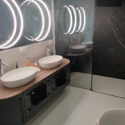 Nowoczesna łazienka z podwójną umywalką nablatową, czarnym blatem z drewnianym akcentem, oświetlonymi okrągłymi lustrami i kabiną prysznicową z czarnymi płytkami imitującymi marmur ze złotymi żyłami.