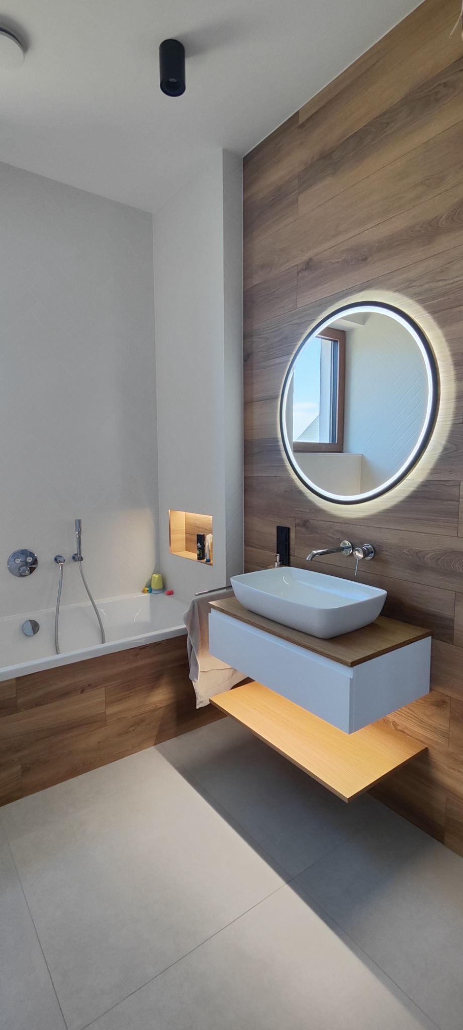 Nowoczesna łazienka z podświetlanym okrągłym lustrem, umywalką nablatową na szafce z podświetleniem, drewnopodobną okładziną ściany i wanną z parawanem.