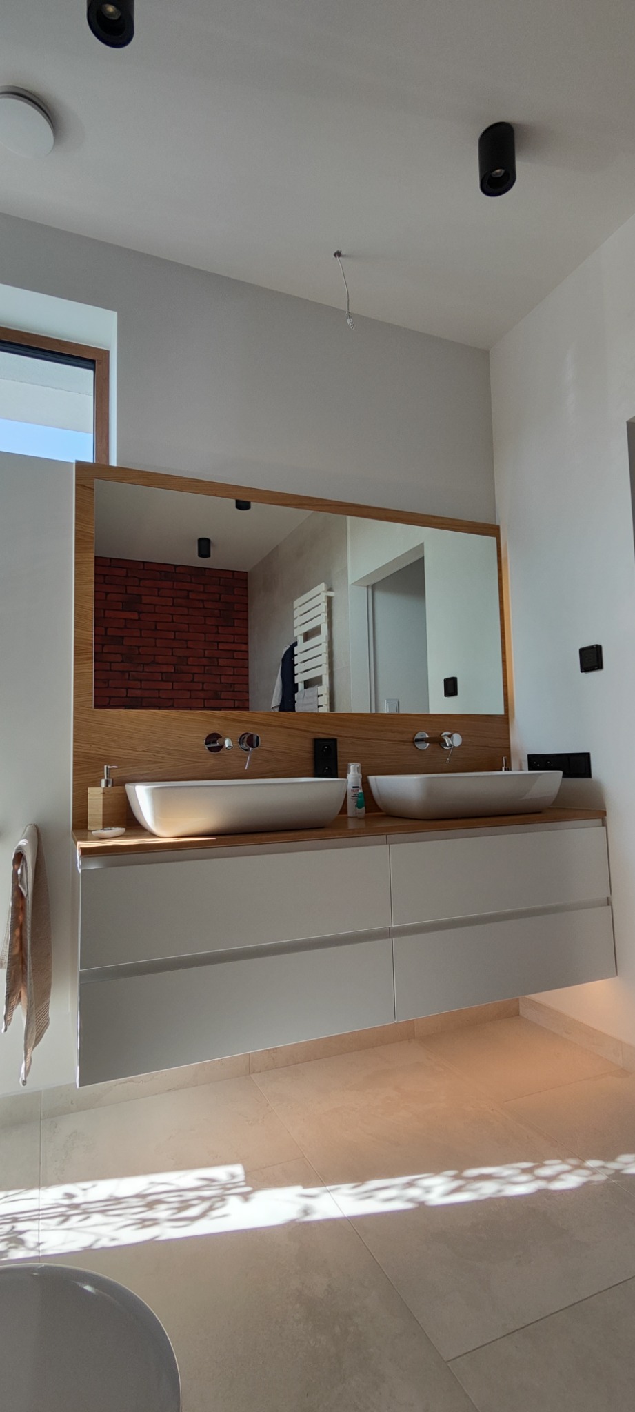 Nowoczesna łazienka z podwójną umywalką nablatową, drewnianym blatem i dużym lustrem, z widoczną ścianą z czerwonej cegły odbijającą się w lustrze.