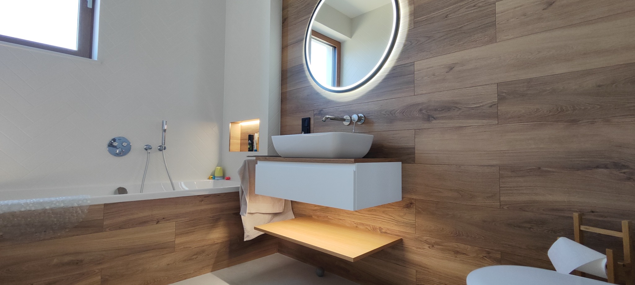 Nowoczesna łazienka z drewnopodobnymi płytkami, okrągłym lustrem z podświetleniem LED, białą umywalką nablatową i wanną obudowaną drewnem.