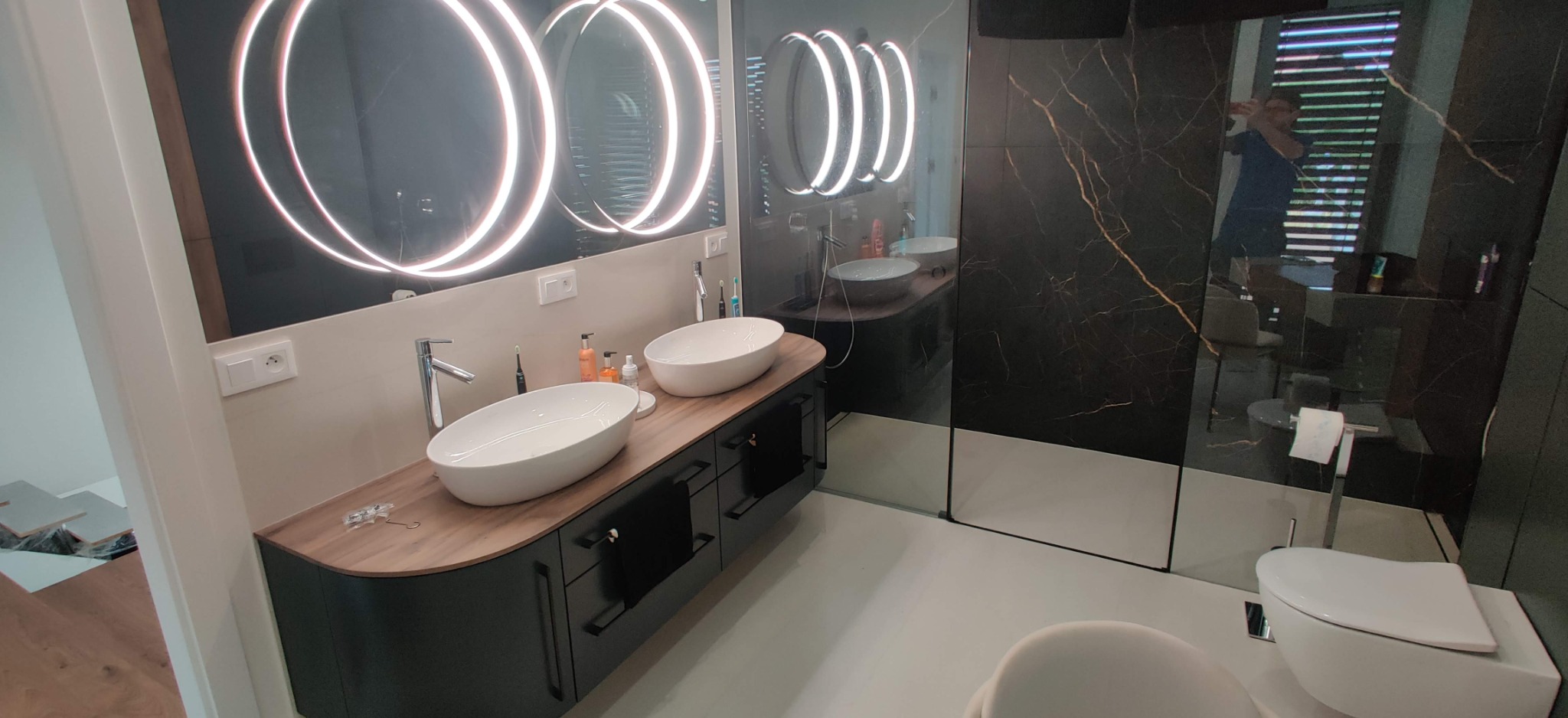 Nowoczesna łazienka z podwójną umywalką nablatową, czarnym blatem z drewnianym akcentem, oświetlonymi okrągłymi lustrami i kabiną prysznicową z czarnymi płytkami imitującymi marmur ze złotymi żyłami.