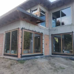 Niedokończony budynek z zamontowanymi oknami przesuwnymi z brązowymi ramami, widoczne cegły i surowy beton, teren wokół budynku nierówny.