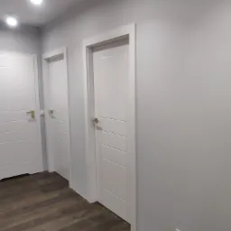 Korytarz z trzema białymi drzwiami wewnętrznymi z poziomymi frezami i złotymi klamkami, oświetlony punktowo, z panelami podłogowymi w odcieniach brązu i szaro-białymi ścianami.