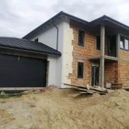 Nowo budowany dom jednorodzinny z zamontowaną, ciemnoszarą bramą garażową segmentową i czarnymi oknami, w trakcie prac wykończeniowych elewacji z widoczną stertą piasku na pierwszym planie.