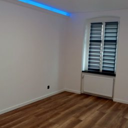 Kris-bud - Puste, odnowione wnętrze pokoju w Legnicy z drewnianą podłogą, białymi ścianami, grzejnikiem pod oknem z roletami, oświetleniem sufitowym i dekoracyjnym niebieskim paskiem LED pod sufitem.