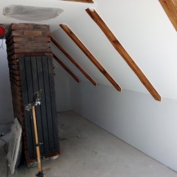 Kris-bud - Poddasze w trakcie remontu: widoczne drewniane belki stropowe, fragment komina z cegły, pomalowane ściany, zakryte folią okno dachowe oraz stojak z narzędziami malarskimi.