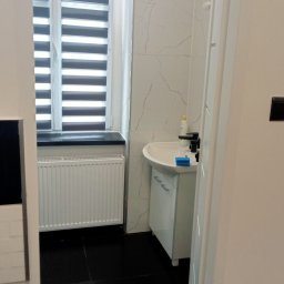 Kris-bud - Umywalka z czarną armaturą w łazience wykończonej białymi płytkami imitującymi marmur, widok zza otwartych drzwi, czarna podłoga z płytek.