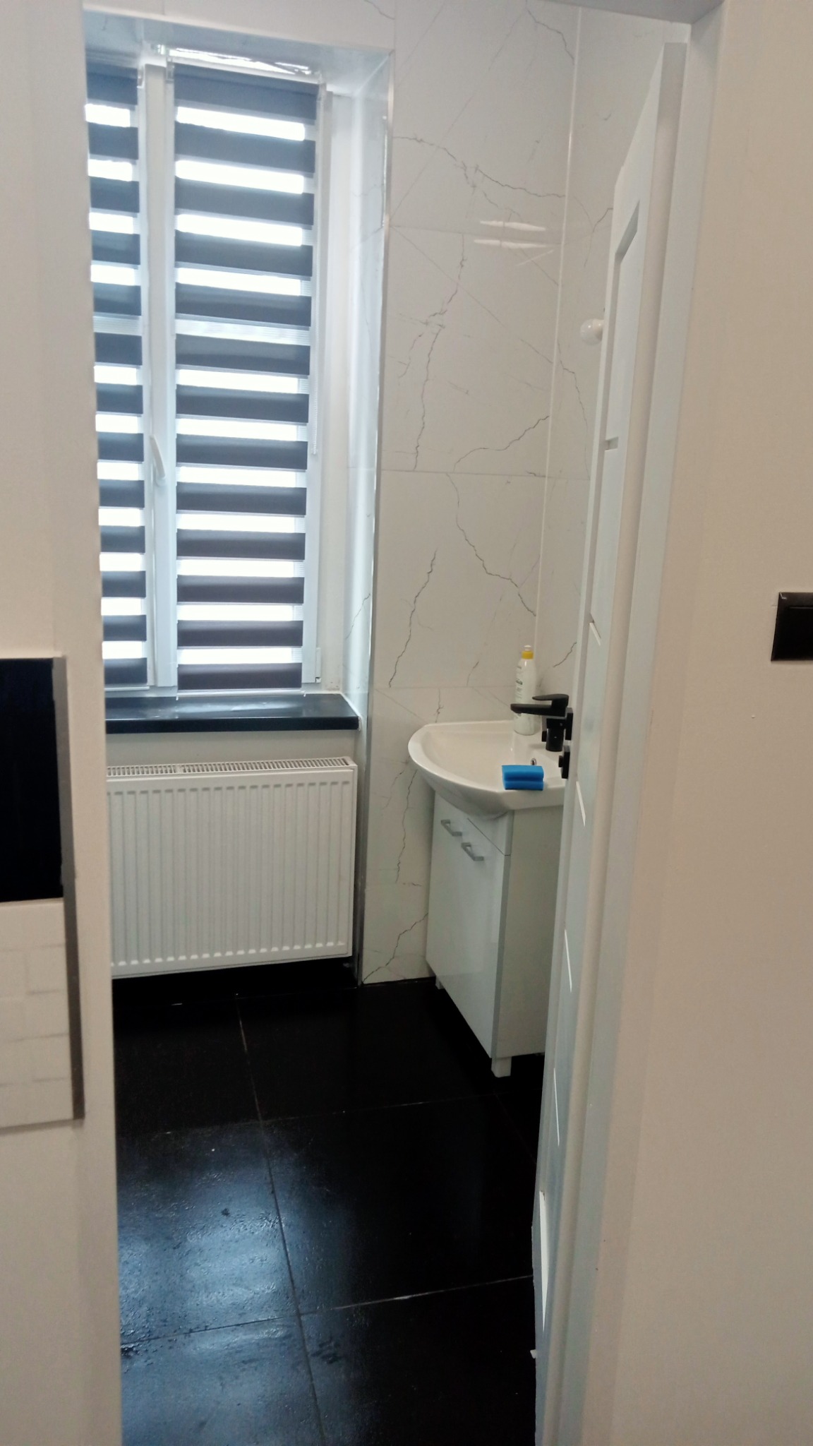 Nowoczesna łazienka z czarną podłogą i białymi marmurkowymi płytkami ściennymi, biała szafka pod umywalką z czarną armaturą, żaluzje okienne dzień-noc.