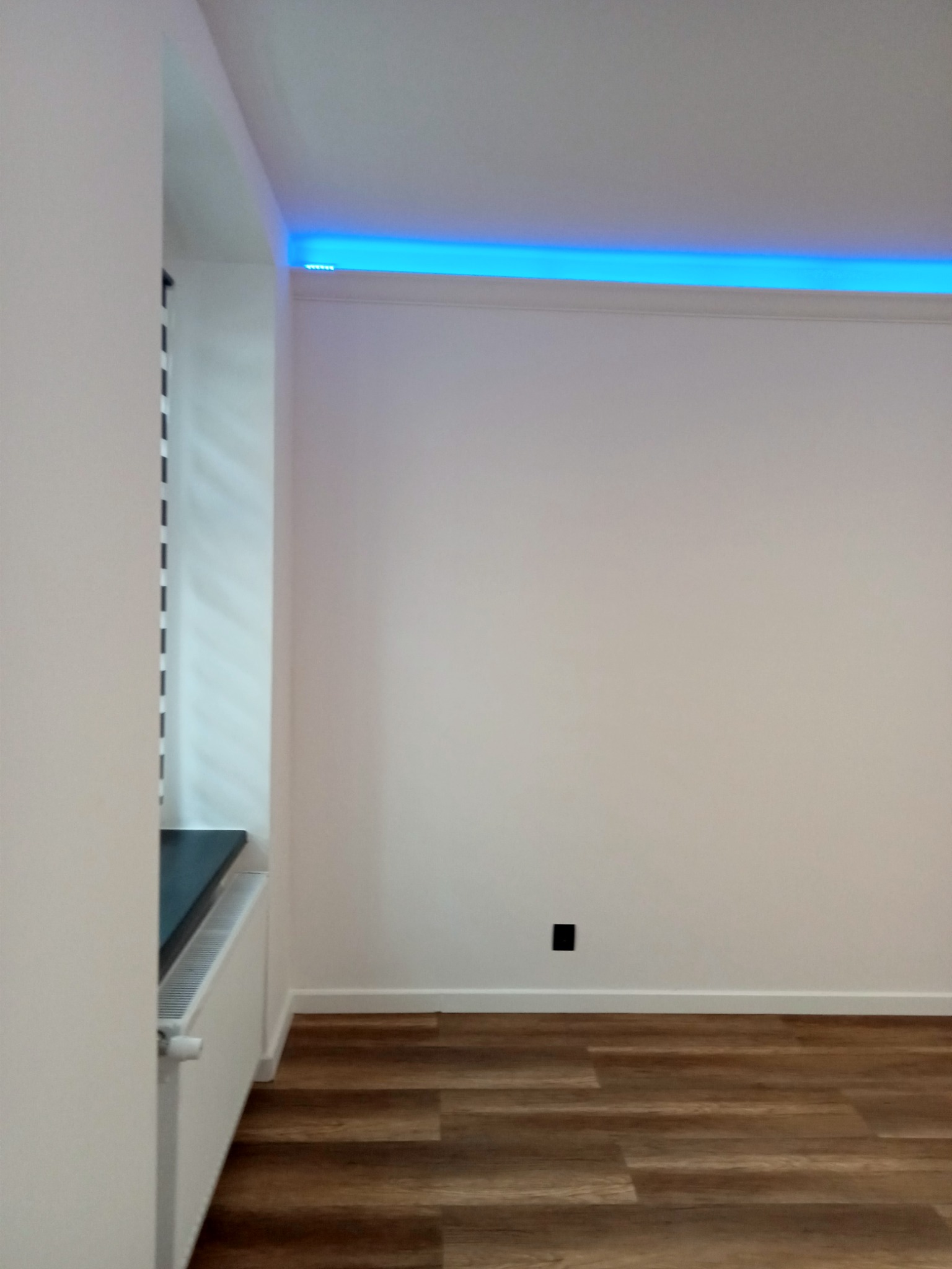 Wnętrze pokoju po remoncie z drewnopodobną podłogą, białe ściany i listwy przypodłogowe, grzejnik pod oknem i niebieskie oświetlenie LED podsufitowe.