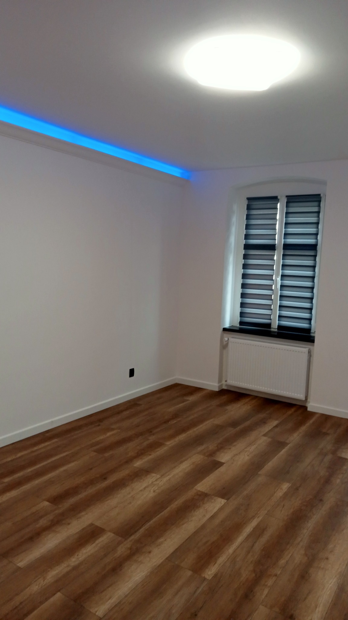Puste, odnowione wnętrze pokoju w Legnicy z drewnianą podłogą, białymi ścianami, grzejnikiem pod oknem z roletami, oświetleniem sufitowym i dekoracyjnym niebieskim paskiem LED pod sufitem.