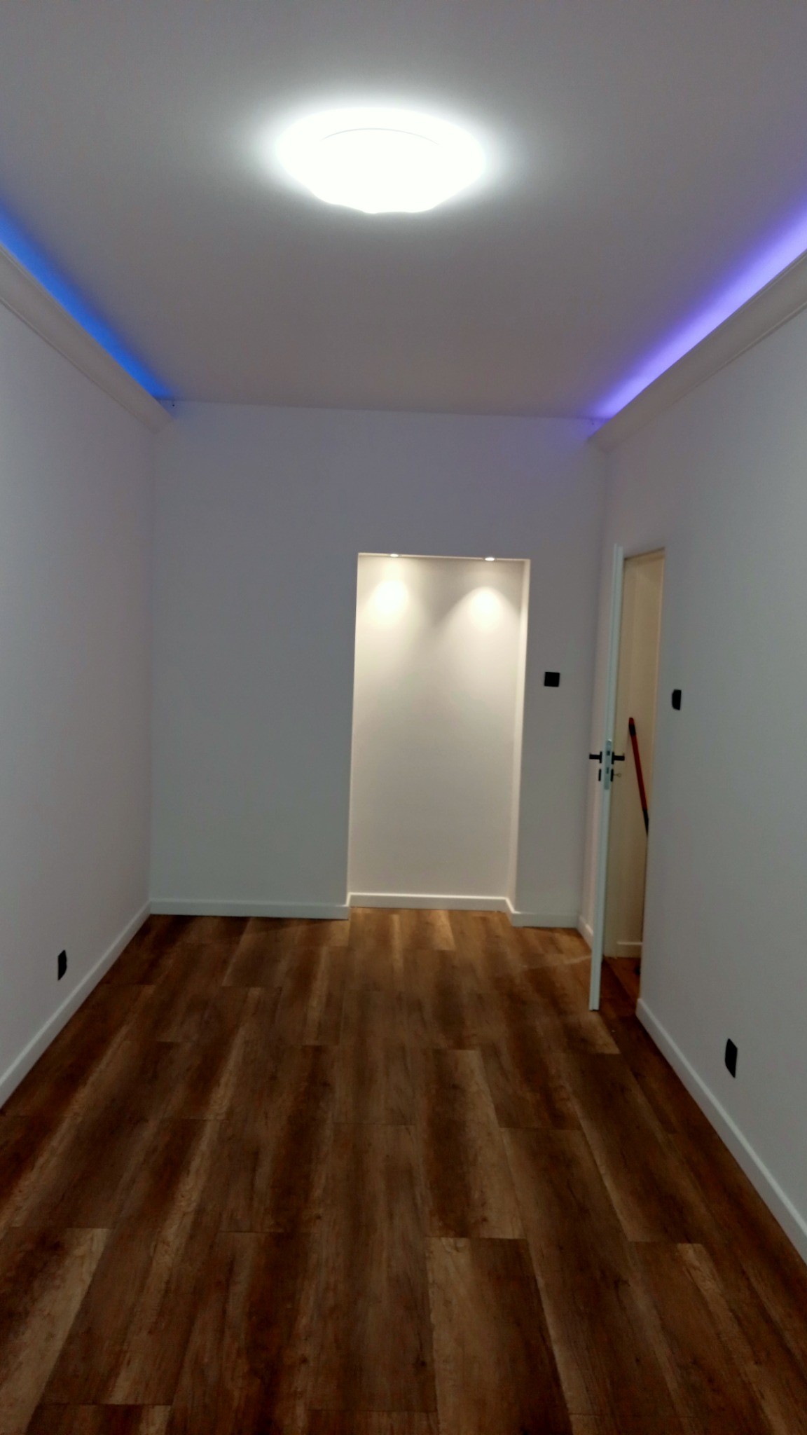 Wnętrze pokoju po remoncie z panelami podłogowymi w odcieniach brązu, białymi ścianami z listwami przypodłogowymi, oświetleniem LED wzdłuż sufitu i wnęką z punktowym oświetleniem.