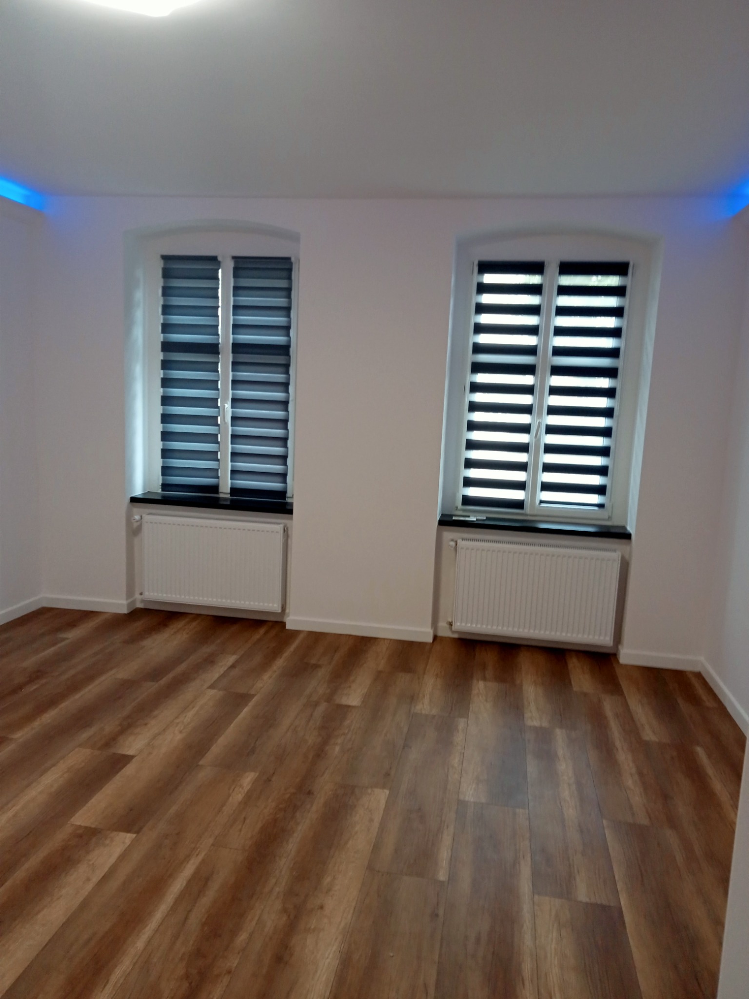 Pusty pokój po remoncie z panelami podłogowymi w kolorze drewna, dwoma oknami z żaluzjami i białymi ścianami z niebieskim oświetleniem LED na suficie.