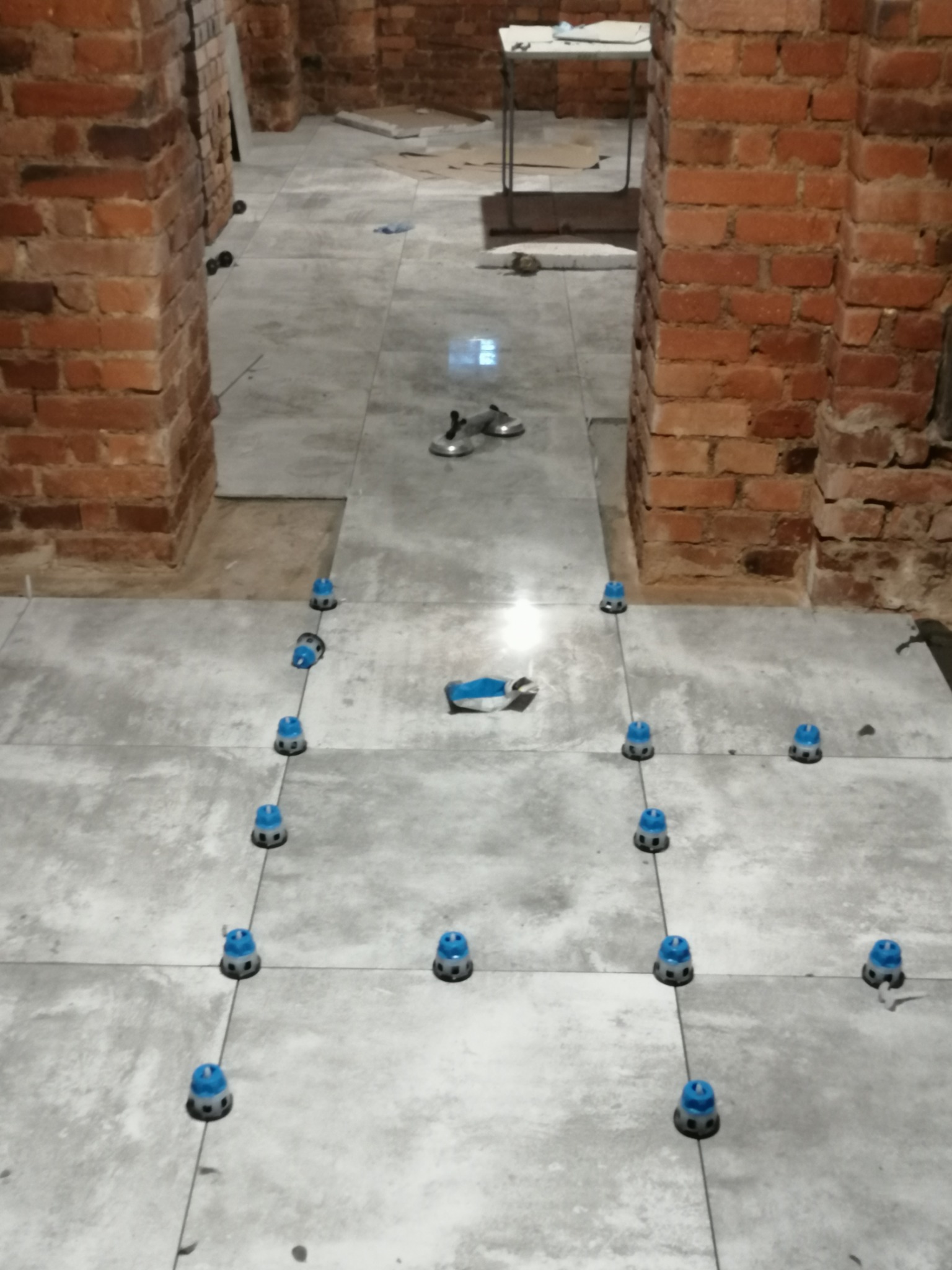 Układanie szarych płytek podłogowych z użyciem niebieskich klipsów poziomujących, widoczne ceglane filary i fragmenty nieułożonych płytek w tle.