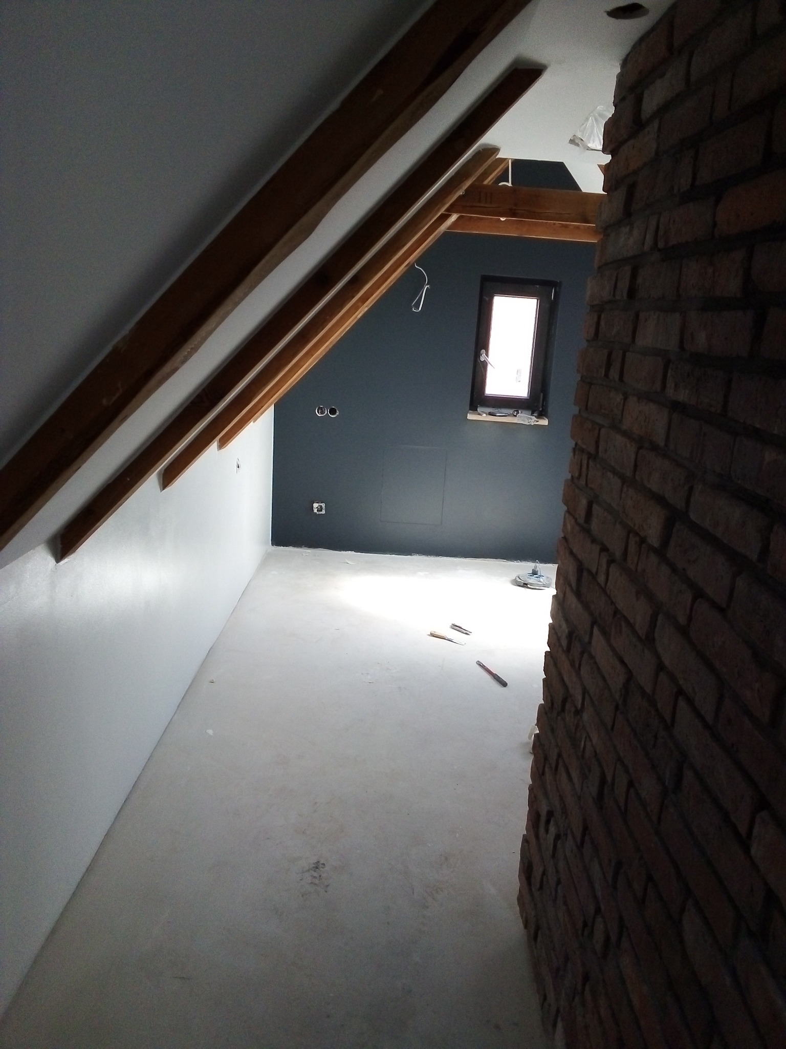 Poddasze w trakcie wykańczania: białe gładzie na skośnej ścianie, surowy beton na podłodze, ciemnoszara ściana z oknem, widoczne drewniane elementy konstrukcyjne dachu i fragment ściany z czerwonej...