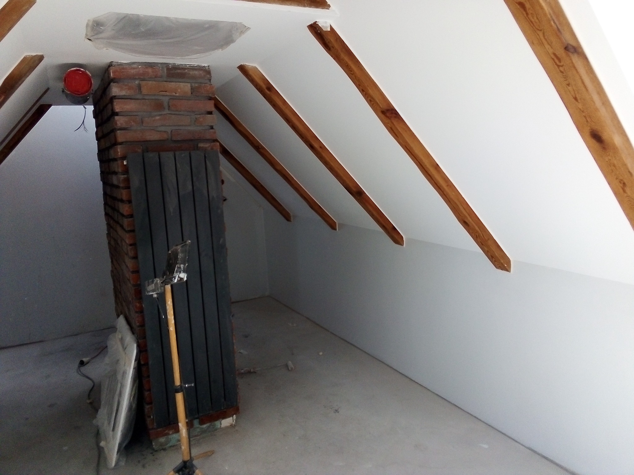Poddasze w trakcie remontu: widoczne drewniane belki stropowe, fragment komina z cegły, pomalowane ściany, zakryte folią okno dachowe oraz stojak z narzędziami malarskimi.